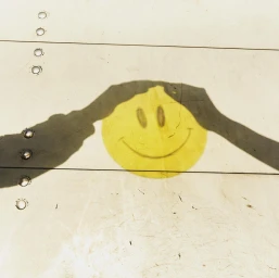 Shadow of human hands framing a yellow painted smiley face on a weathered white metallic surface with rivets