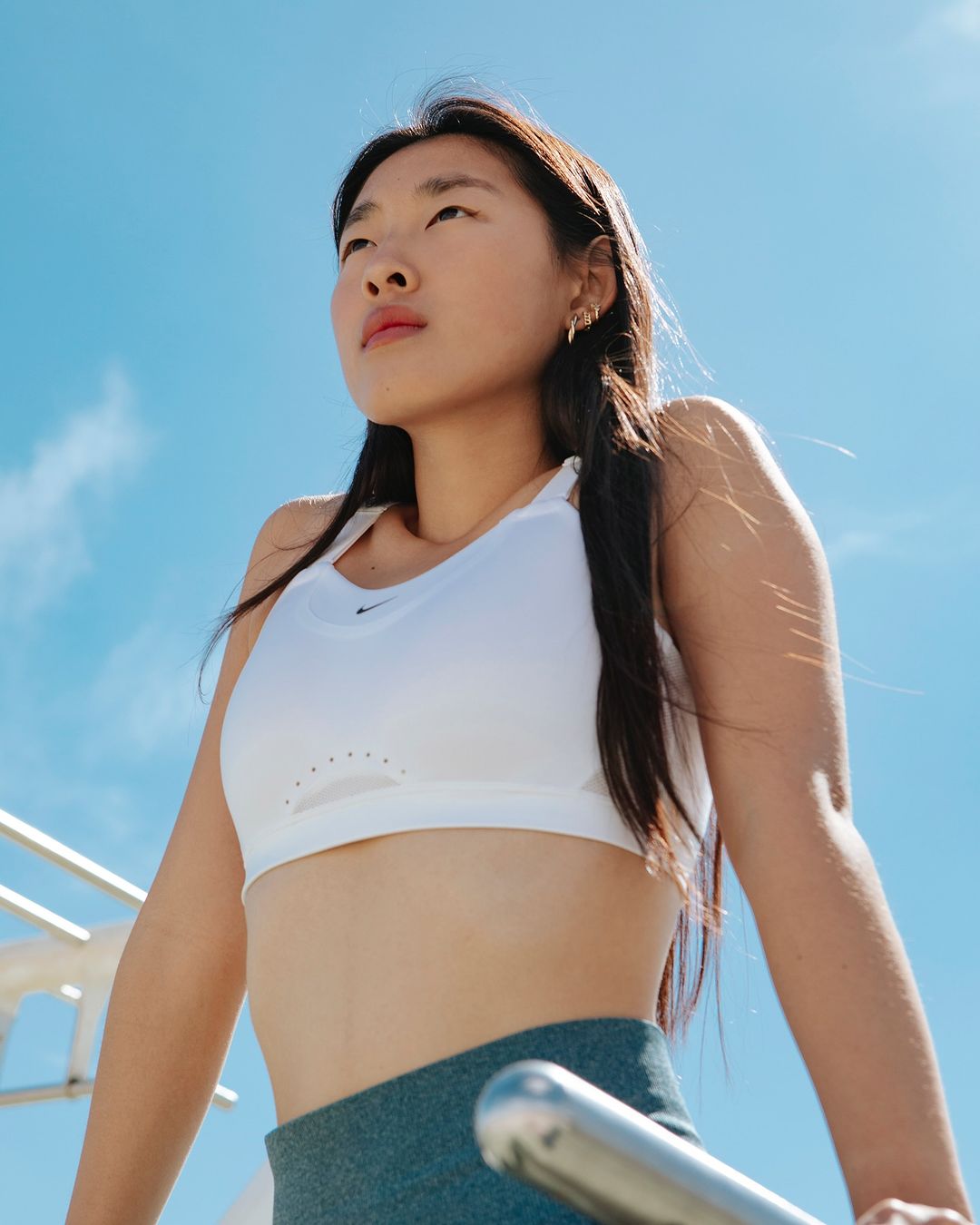 East Asian woman in a white sports crop top and slate blue leggings against a clear blue sky by Rocio Chacon
