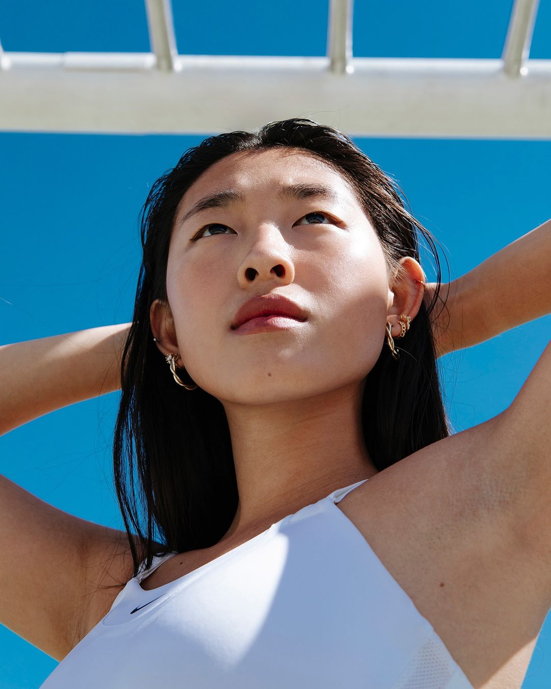 Low-angle shot of a young Asian woman in a white top, looking upwards with a serene expression against a bright blue sky and white bars.