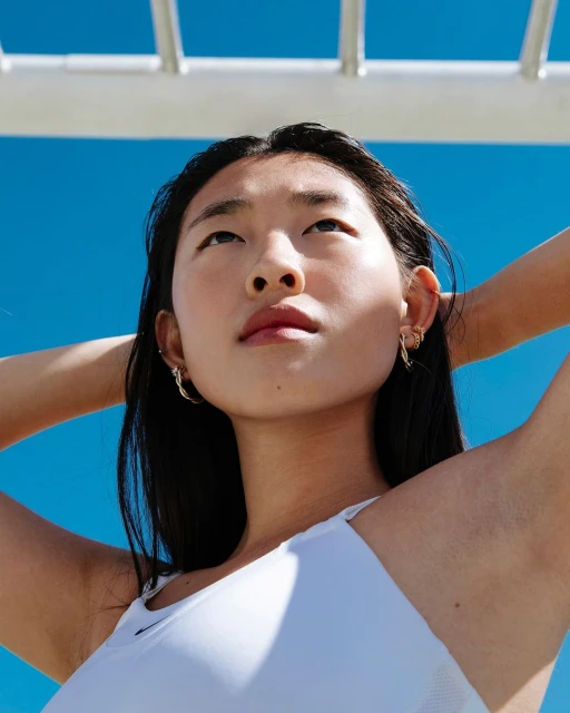 Low-angle shot of a young Asian woman in a white top, looking upwards with a serene expression against a bright blue sky and white bars.