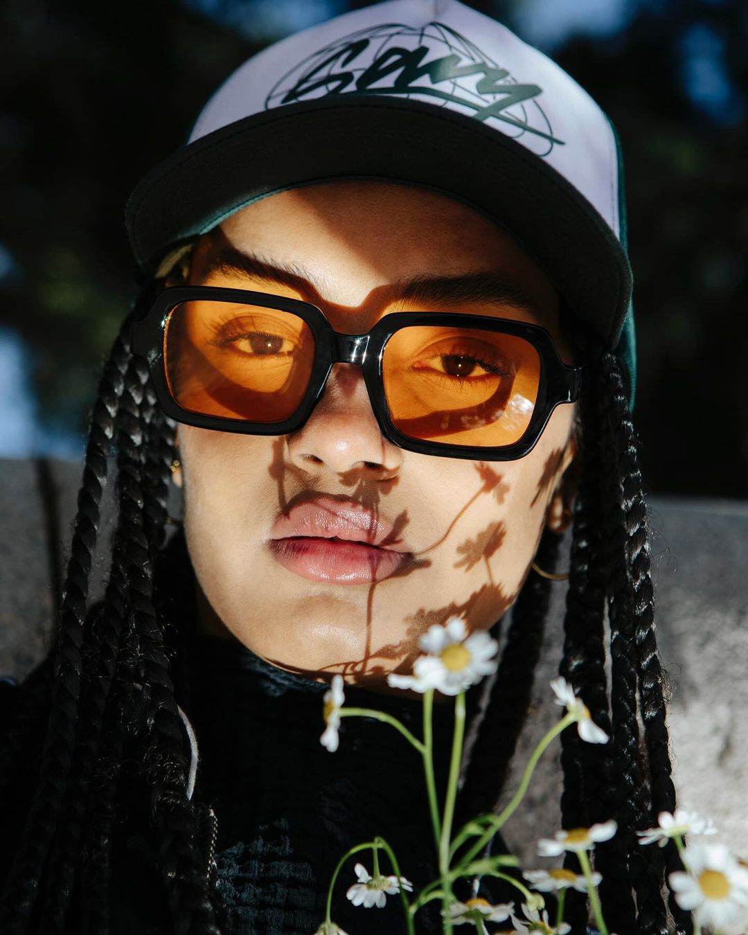 Woman poses with a baseball cap and orange glasses in a close-up shot with daisy flowers and shadows across her face.