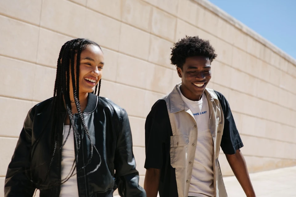Two smiling young adults walk outdoors against a neutral wall. The woman wears braids and a black jacket. The man wears a two-tone shirt.