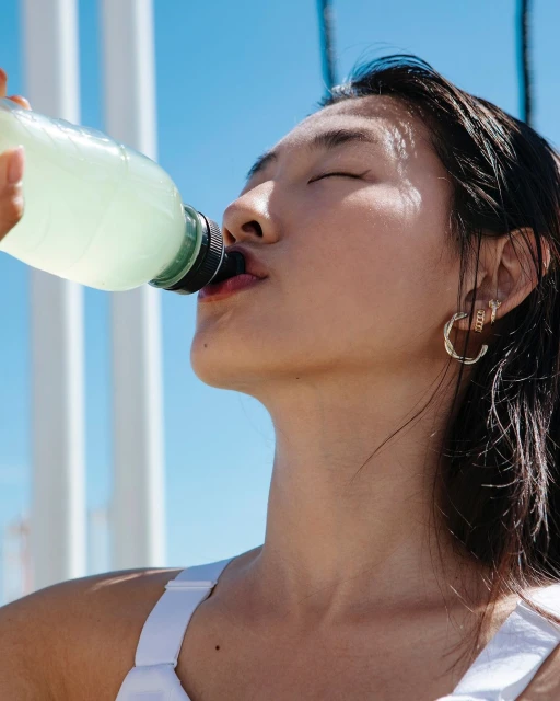 Close-up of a young woman drinking from a clear bottle in front of a blue sky and white architectural structure on a sunny day.