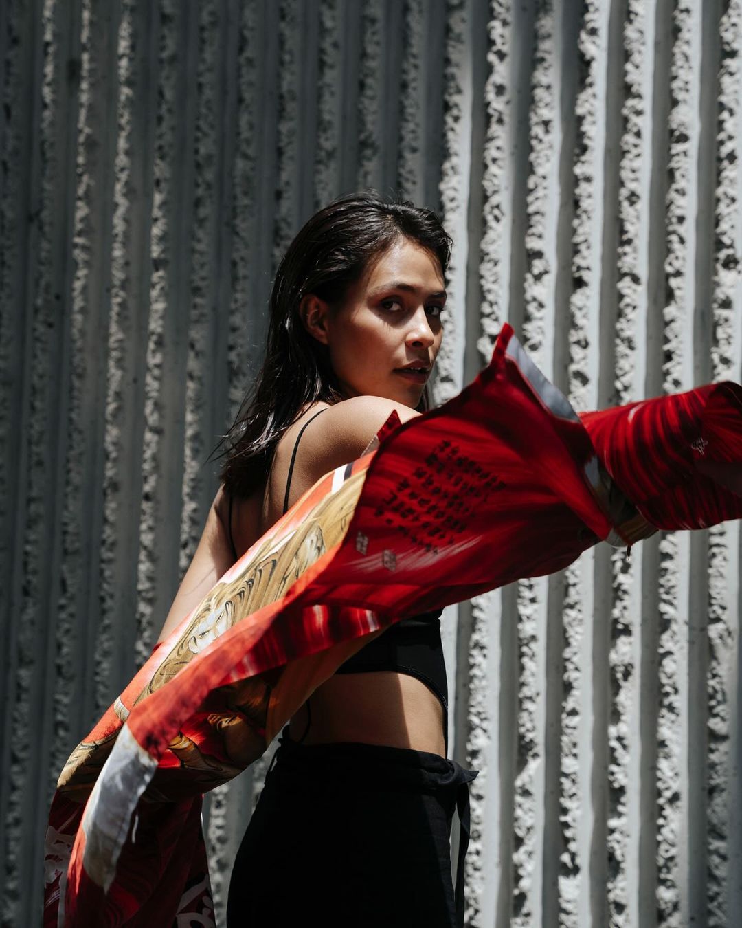 A woman in a black top and pants stands in front of a textured wall, holding a red patterned scarf. She glances over her shoulder.