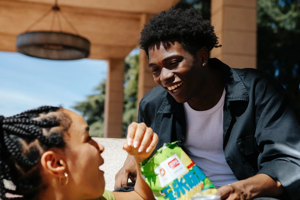 Two young adults are laughing while sharing a bag of chips on a sunny day, with a stone building and trees in the background.