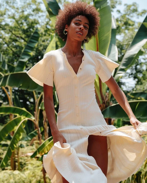 A woman models a cream-colored dress with flutter sleeves outdoors, showcasing a blend of fashion and natural background.