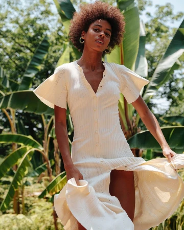 A woman models a cream-colored dress with flutter sleeves outdoors, showcasing a blend of fashion and natural background.