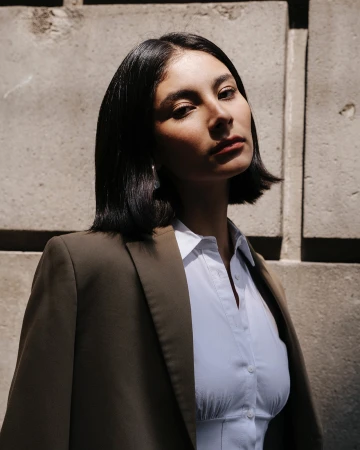 Portrait of a woman with short dark hair wearing a white shirt and olive blazer against a textured concrete wall.