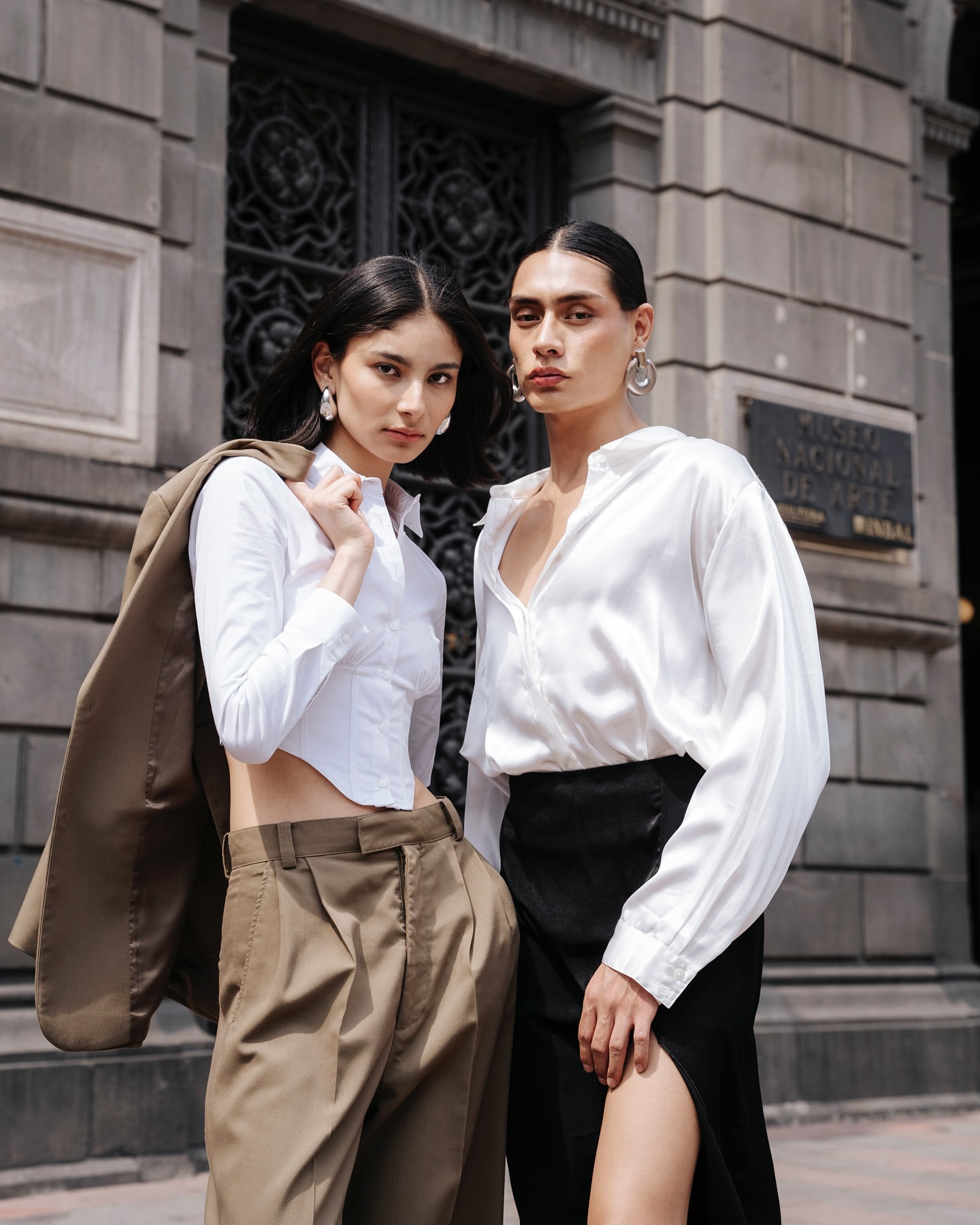 Two fashion models pose in front of a stone building; one in brown slacks and a white blouse, the other in a black skirt and blouse.