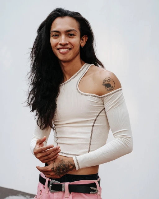 A smiling adult with long dark hair wears a cream long-sleeve shirt and pink pants in a medium shot against a neutral background.