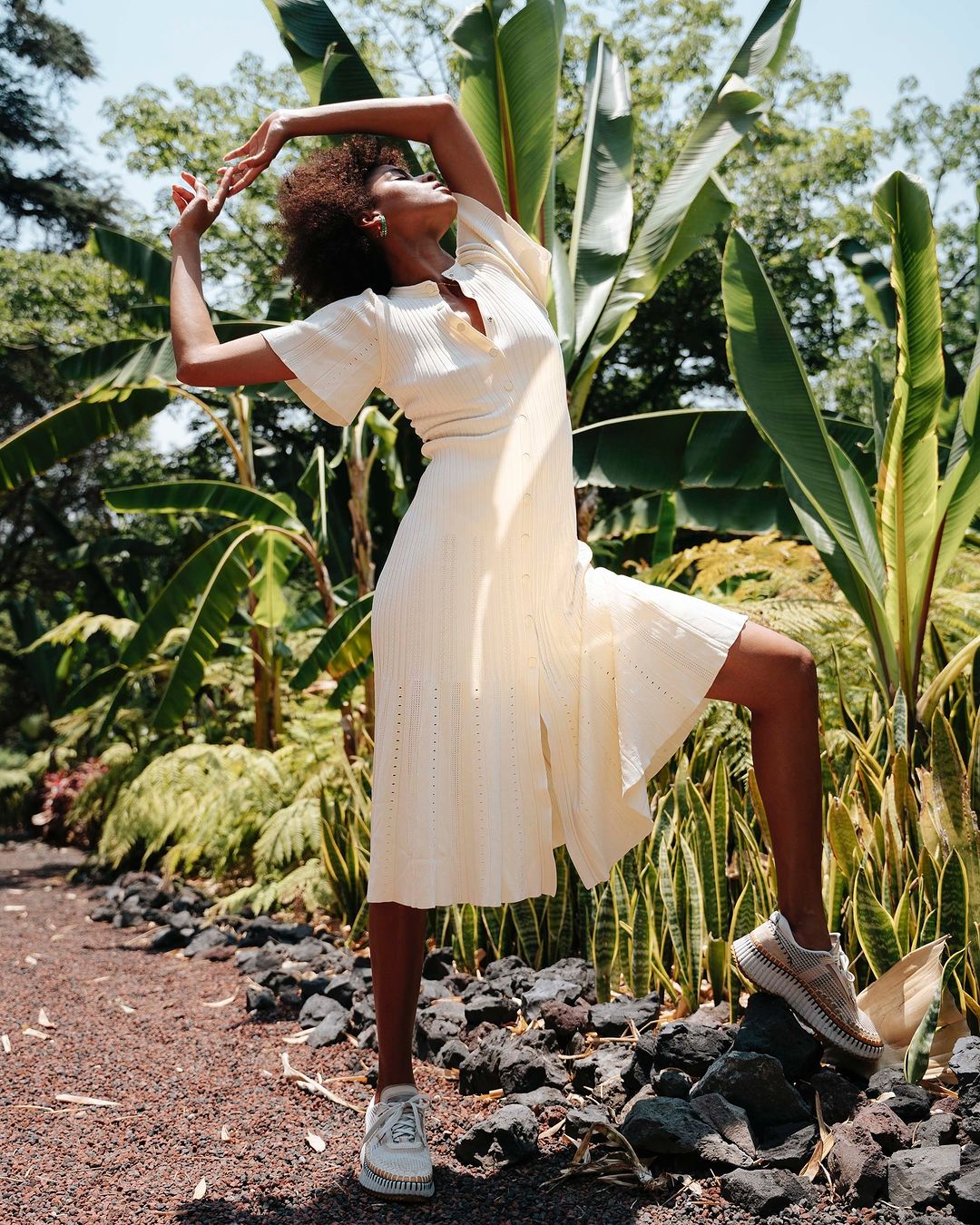 A woman poses in a cream dress and sneakers among lush tropical foliage and rocks in an outdoor setting, bathed in bright daylight.