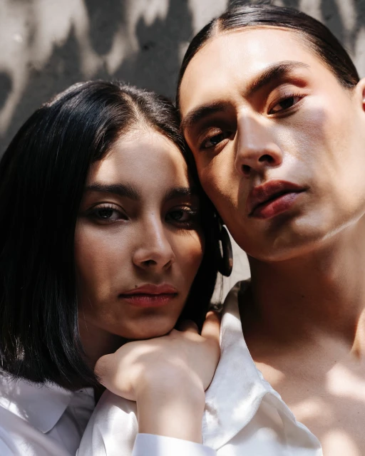 Close-up portrait of a woman resting her head on the shoulder of another person, both wearing white shirts and illuminated by dappled sunlight.
