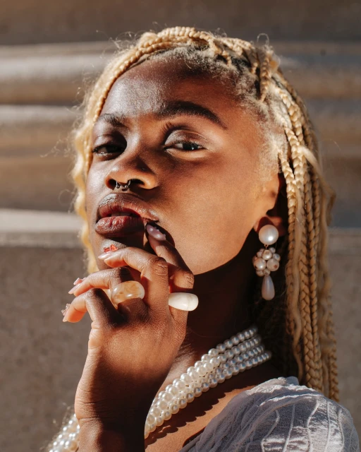 Close-up portrait of an African-descent woman with blonde braided hair, wearing pearl jewelry, and posing in front of a wall.