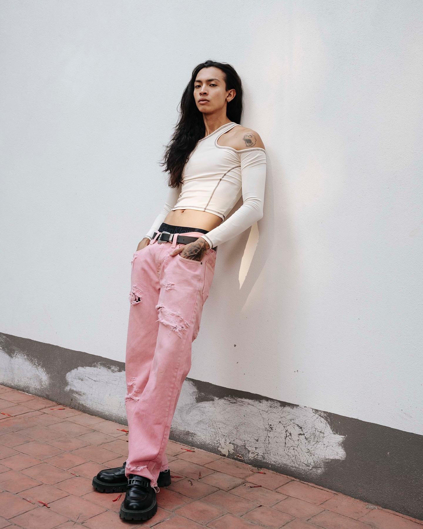 A slender adult male with long hair leans against a white wall, wearing a beige top, ripped pink jeans, and black loafers.