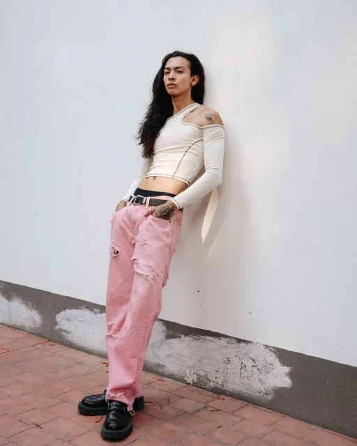 A slender adult male with long hair leans against a white wall, wearing a beige top, ripped pink jeans, and black loafers.