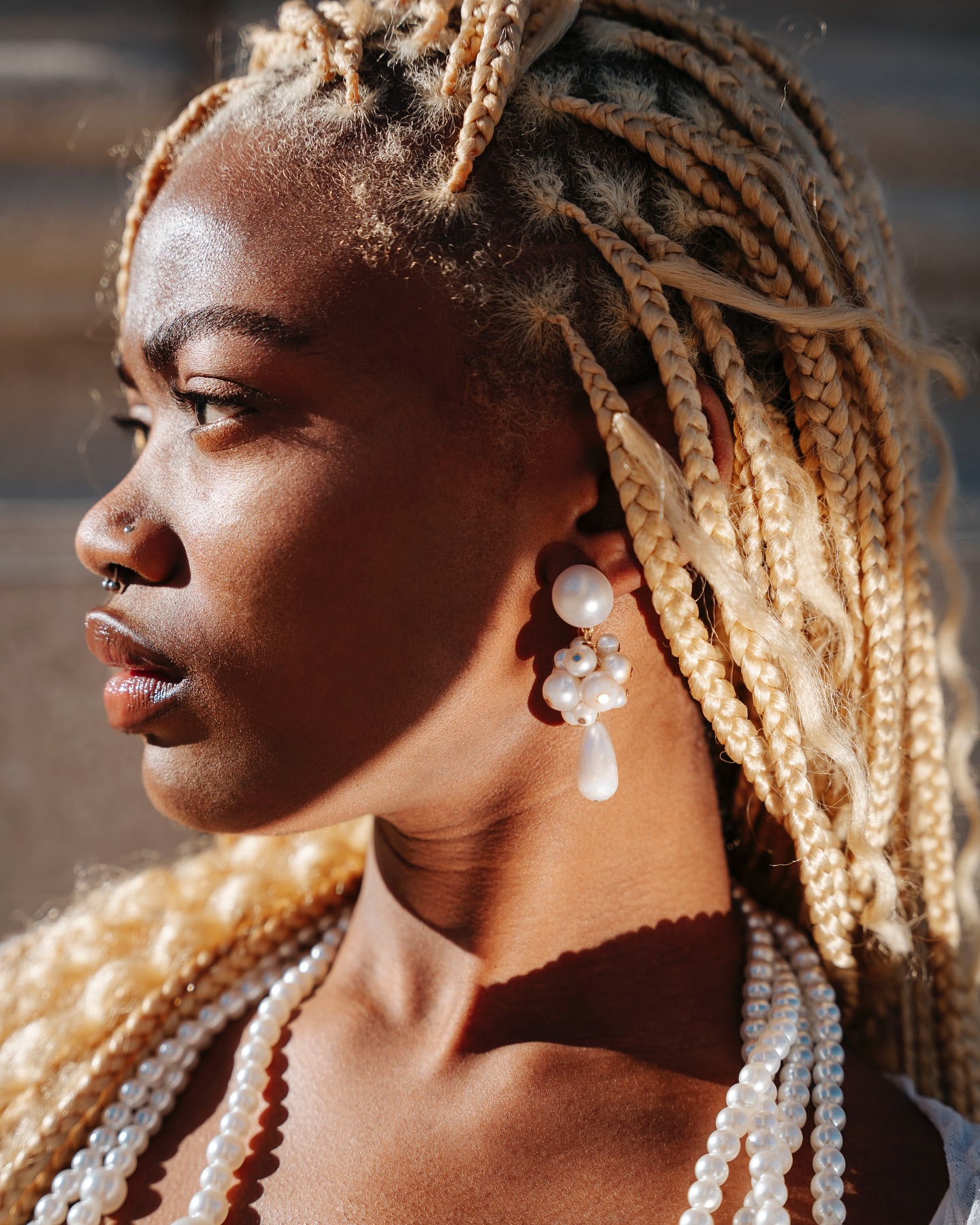 Close-up of a Black woman with blonde box braids wearing pearl earrings and a pearl necklace, illuminated by natural sunlight.
