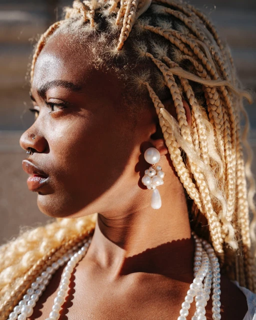 Close-up of a Black woman with blonde box braids wearing pearl earrings and a pearl necklace, illuminated by natural sunlight.