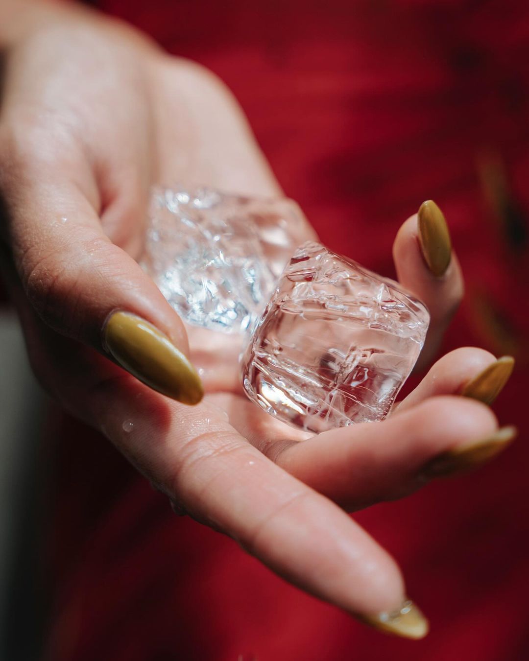 Close-up of a hand with gold nails holding three clear ice cubes against a blurred red fabric background. Focus on the cubes.