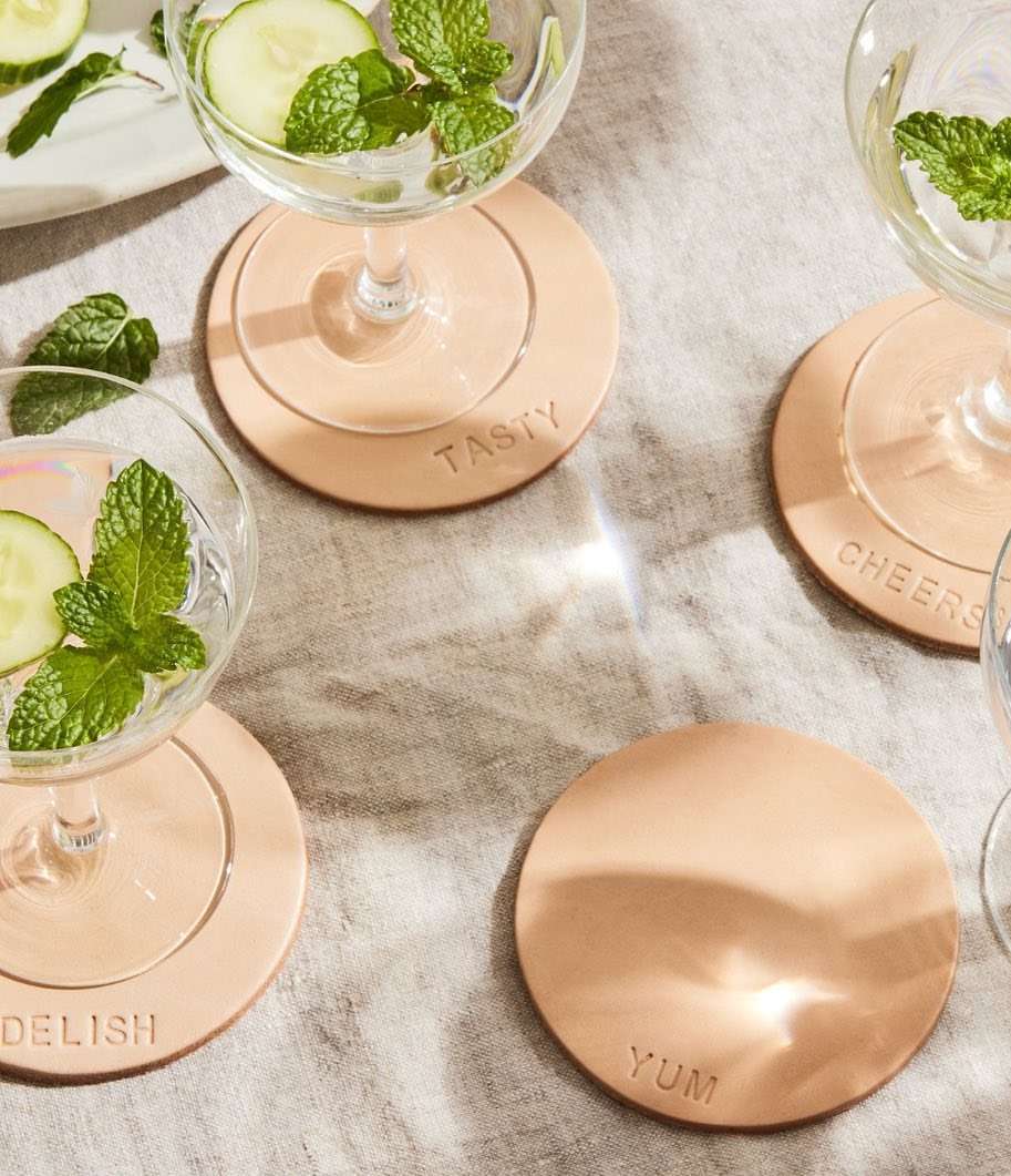 Overhead shot of several cocktail glasses filled with clear beverages, cucumber slices, and mint, resting on tan leather coasters.