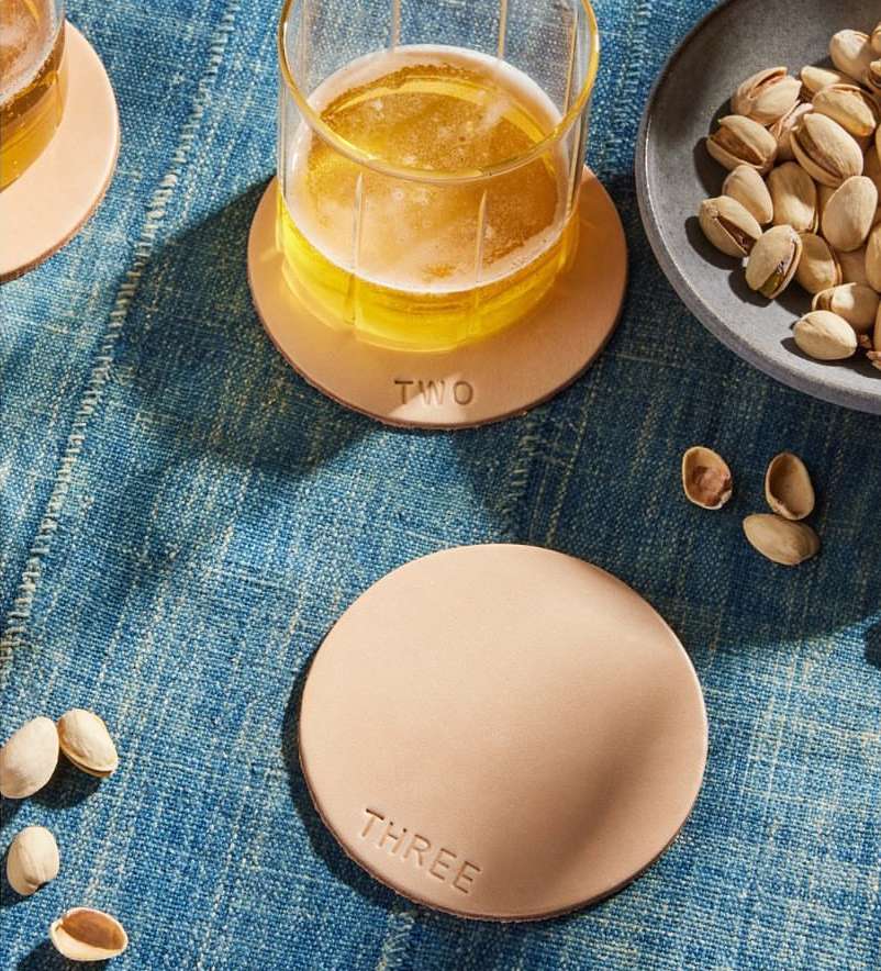 Close-up of a beer in a glass on a leather coaster with pistachios and a blue patterned tablecloth, offering a refreshing and inviting scene.