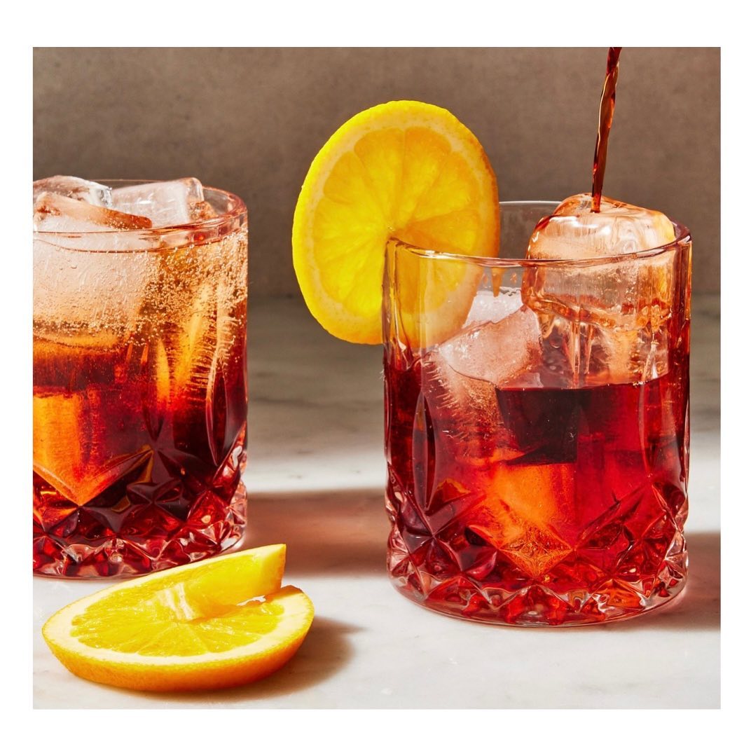 Two crystal glasses of deep red cocktail with ice cubes and orange garnish, beverage being poured in, neutral background. Food photography.