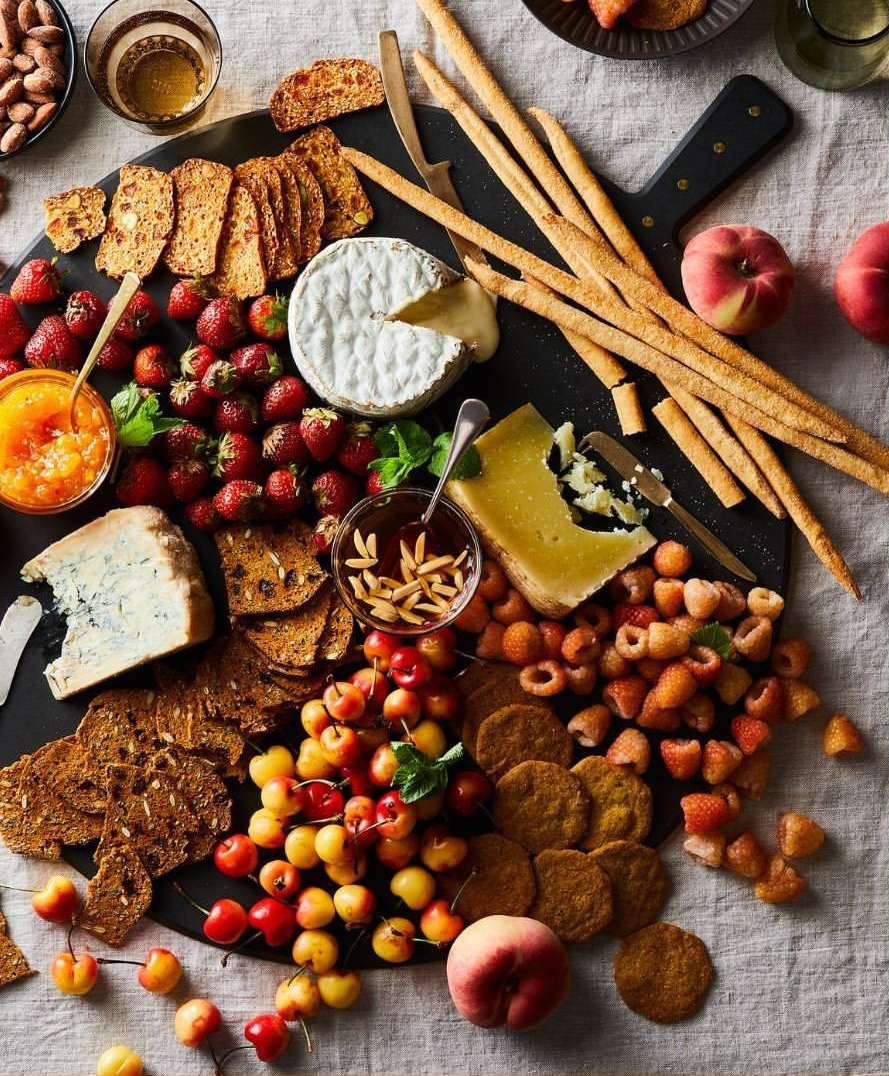 Overhead shot of a food board with various cheeses, strawberries, cherries, raspberries, crackers, fruit preserves, and breadsticks.
