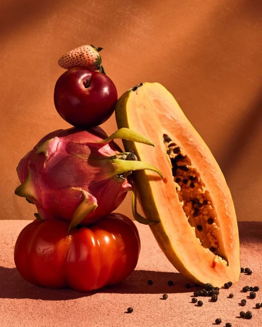 Still life of a stacked tomato, dragon fruit, plum, and strawberry next to a halved papaya, creating a vibrant display of fresh produce.
