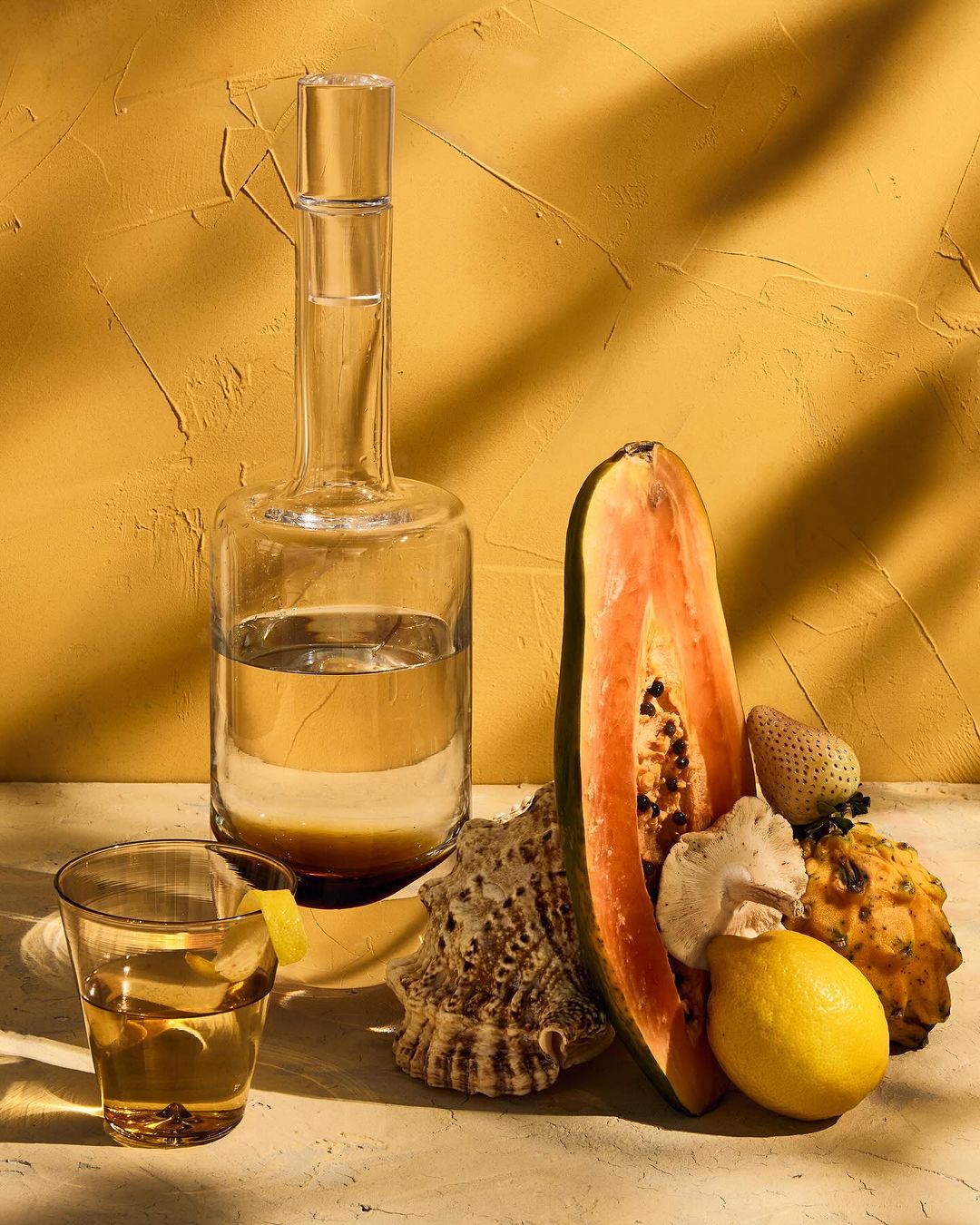 Still life with a glass bottle, a drink in a glass, papaya half, lemon, and seashells arranged against a textured yellow background.