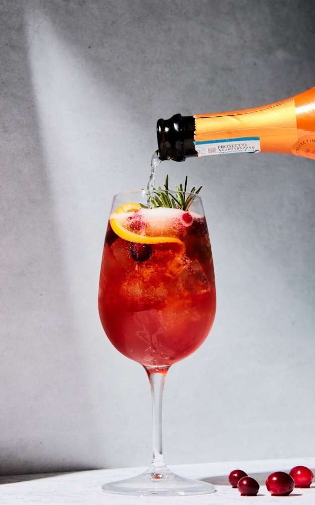 Sparkling wine being poured into a stemmed glass with cranberries, orange peel, and rosemary, set against a textured gray backdrop.
