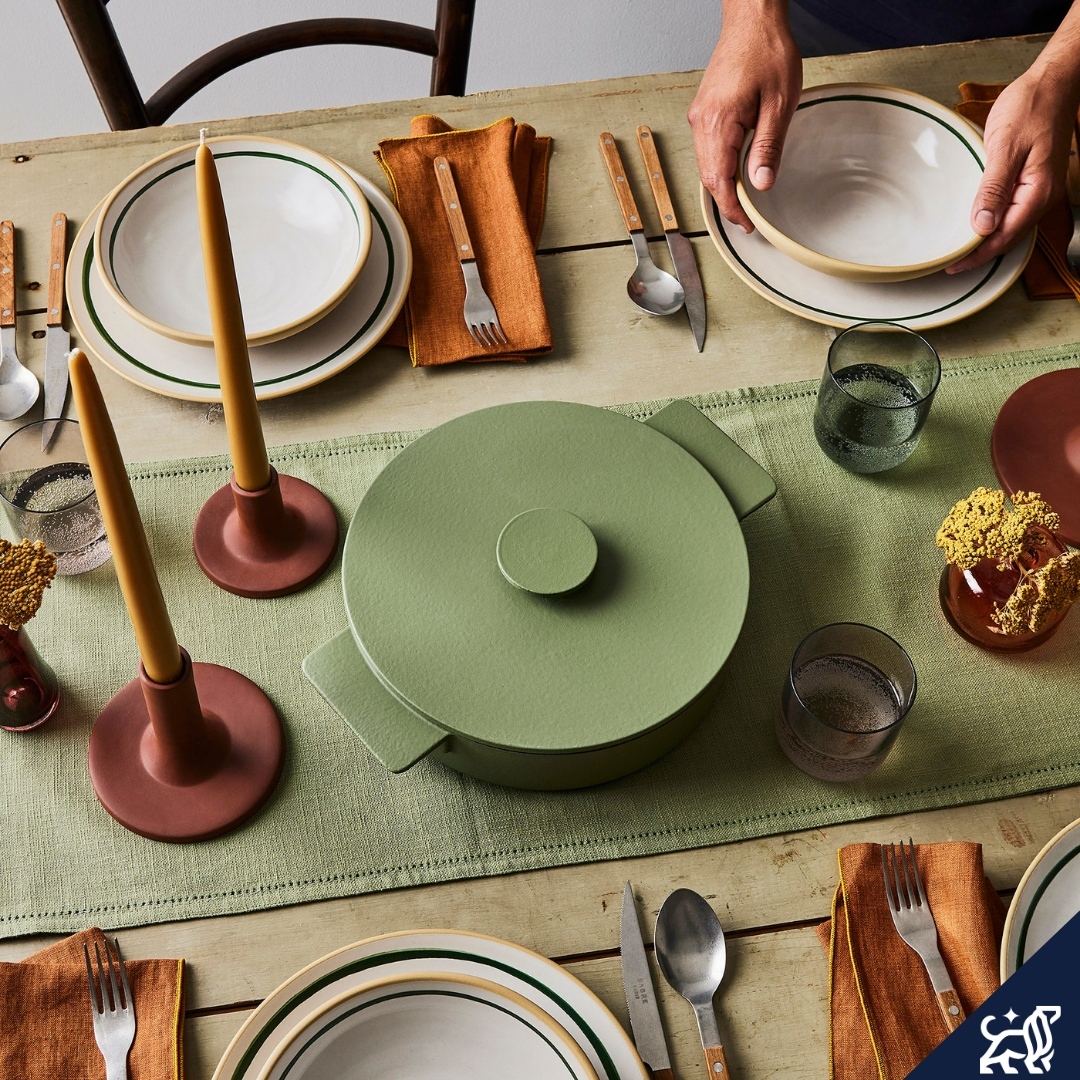 Overhead shot of a rustic wooden table setting with plates, cutlery, green pot with lid, candles, and glassware. Neutral tones.