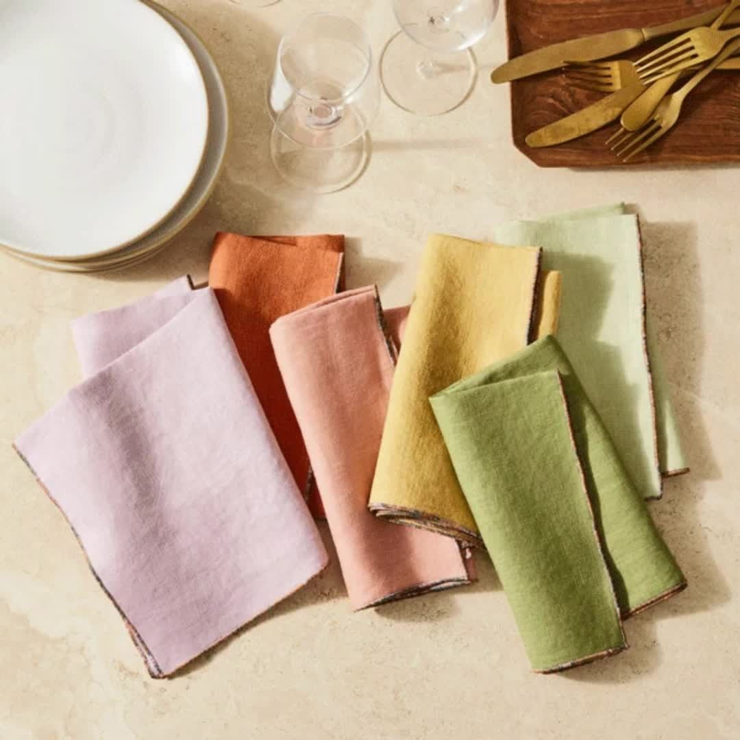 Overhead shot of pastel-colored linen napkins neatly arranged on a neutral surface with plates, glassware, and gold cutlery.
