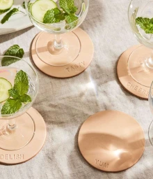 Glassware with cucumber slices and mint leaves sitting on round leather coasters with embossed text over linen.