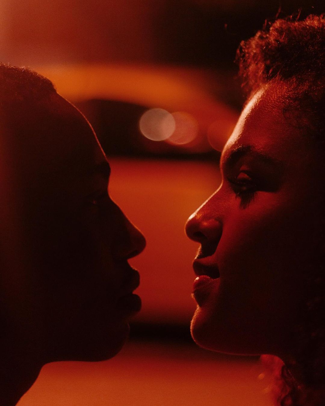 Close-up profile of a man and a woman in red lighting, faces near each other, creating an intimate and warm atmosphere.