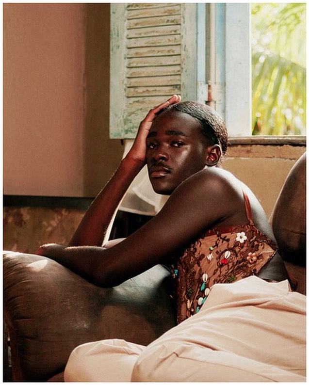 Indoor portrait of an African American person sitting on a sofa in front of a window with white shutters, looking at the camera.