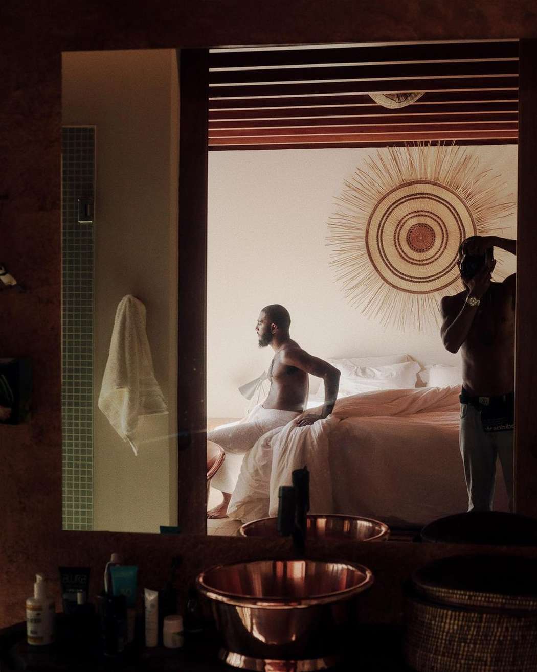 Reflection in a bathroom mirror shows two Black men in a bedroom, one sitting on a bed and the other taking a photo with a camera.