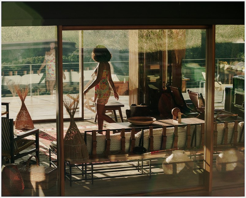 A woman in a floral dress walking on a wooden deck with glass sliding doors leading to a warmly lit interior and outdoor greenery.