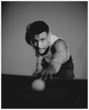 Monochrome portrait of a man with a ball, aiming at a billiards table in a photography studio setting with soft, even lighting.