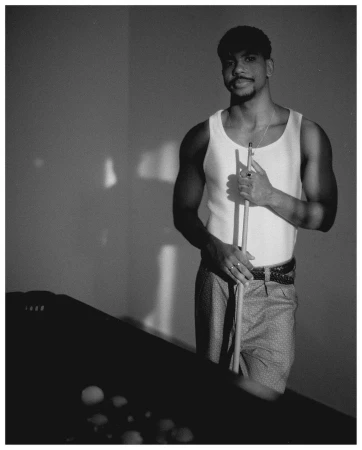 Monochrome portrait of a man in a tank top holding a pool cue next to a billiard table indoors with natural lighting from the side.