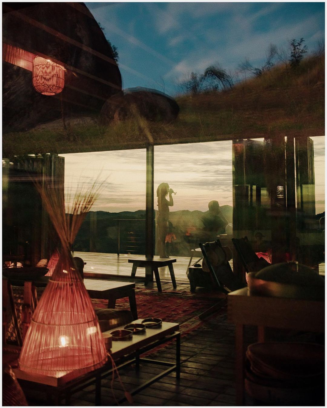 Indoor view through glass doors reveals woman silhouetted at dusk. Lanterns glow in the room with natural outdoor landscape beyond.