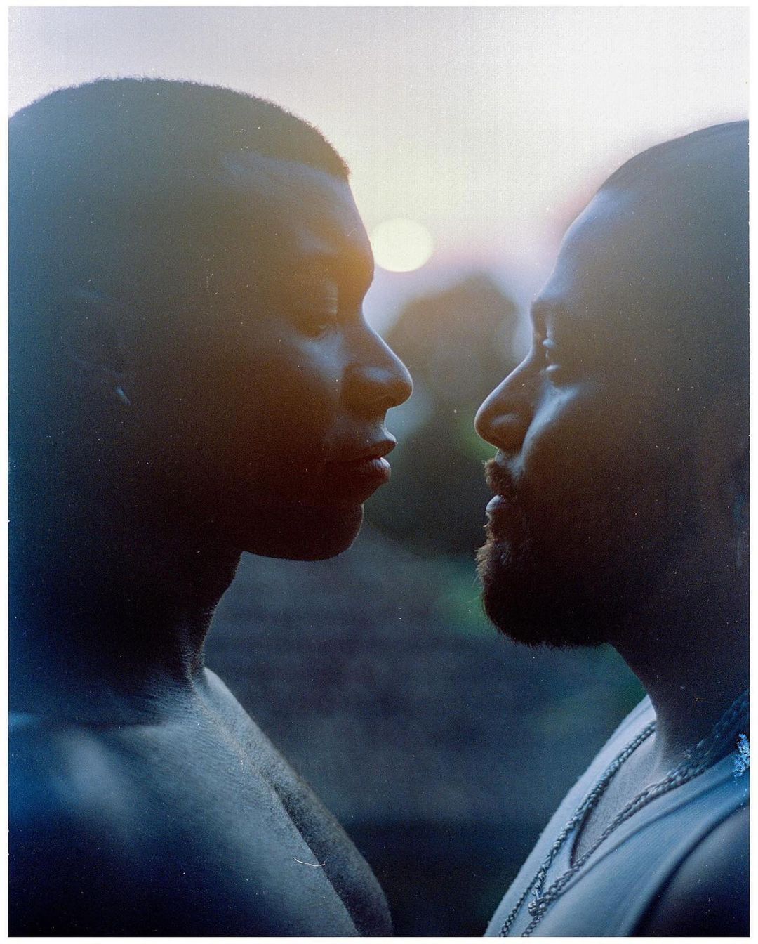 Close-up portrait of two adult men, face to face, with blurred background and sunlight, one wearing a chain and tank top.