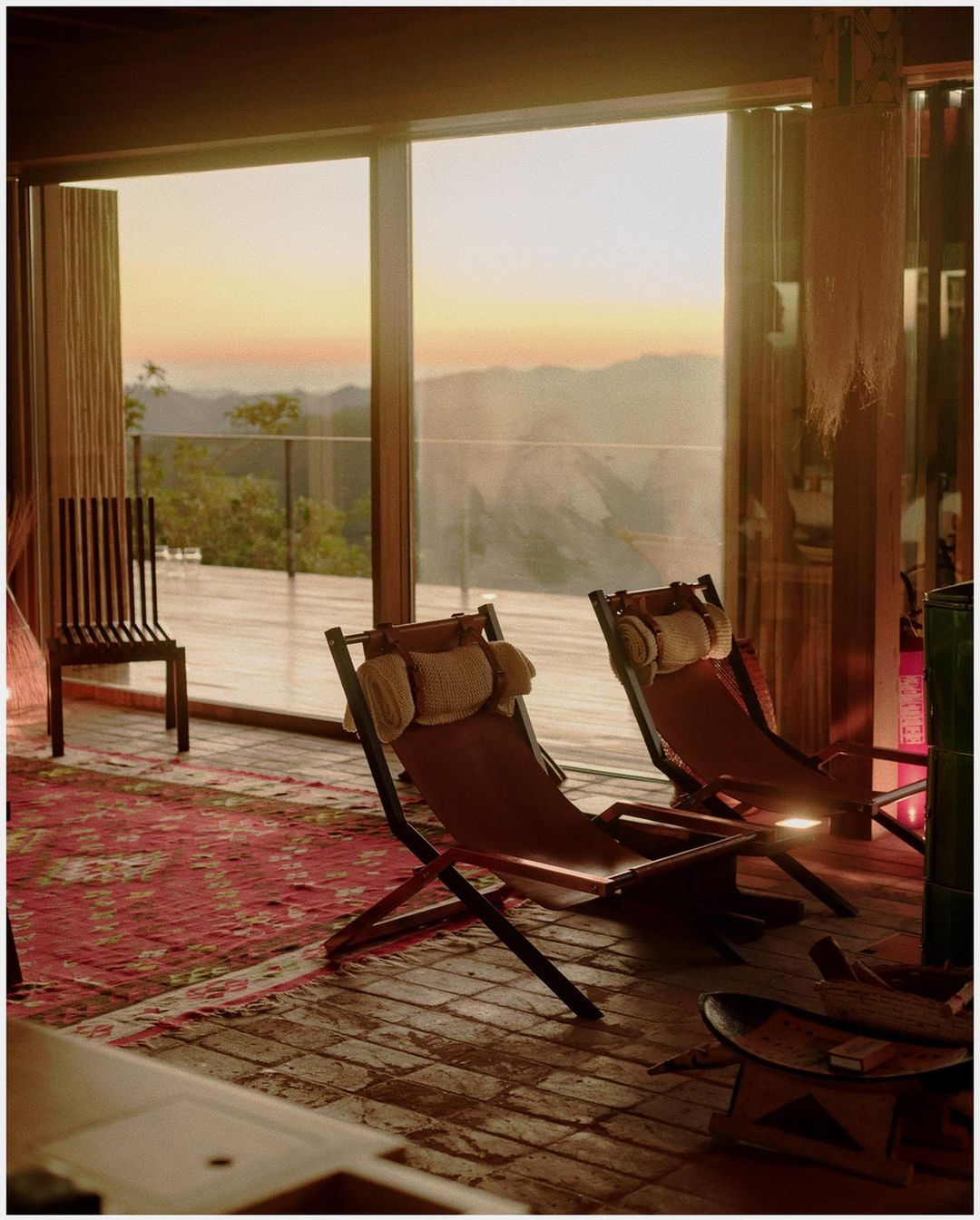 Interior view with two tan canvas sling chairs, wooden floors and a patterned red rug, looking out through windows to a mountain sunset.