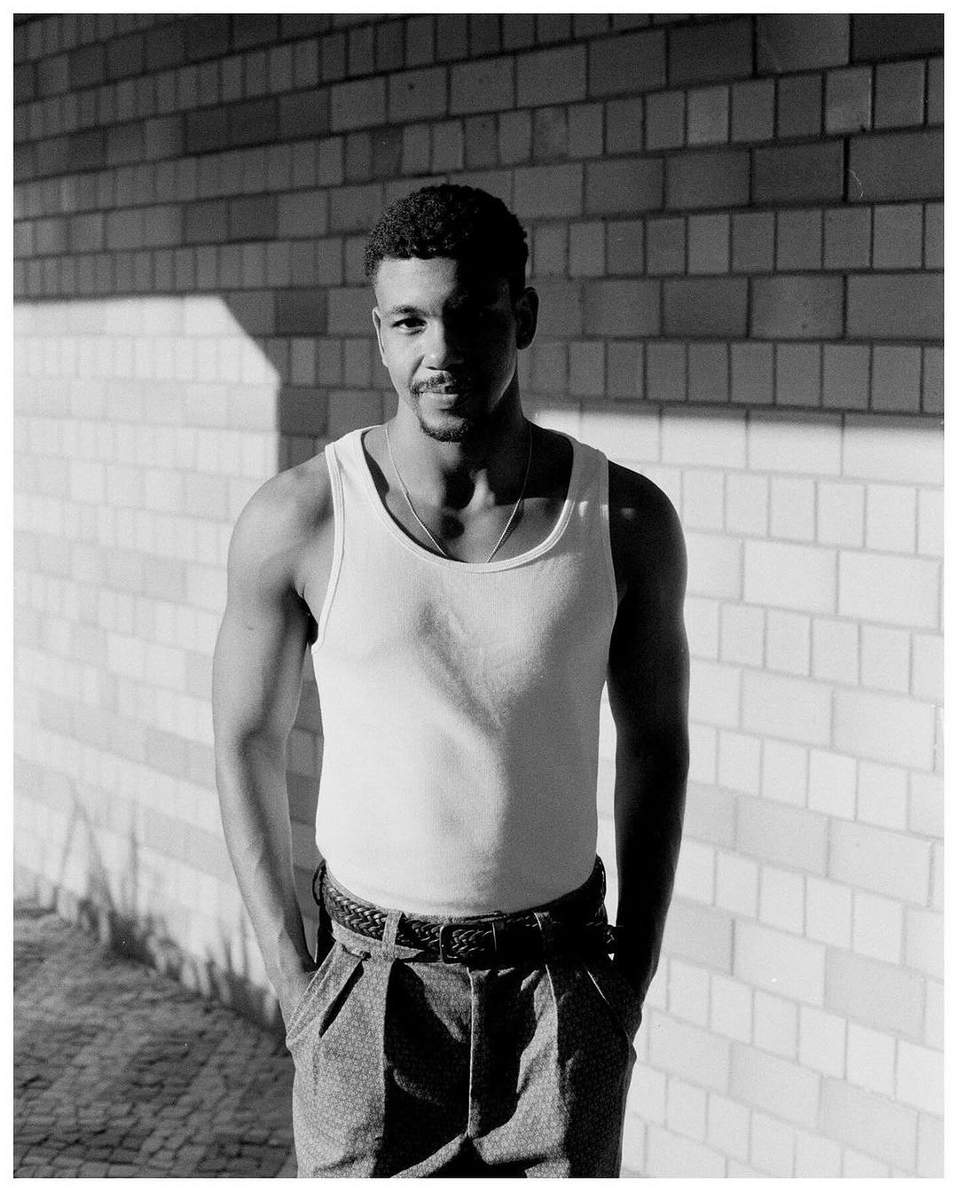Portrait of a Black man in a white tank top standing against a brick wall, photographed by Rodrigo Oliveira.