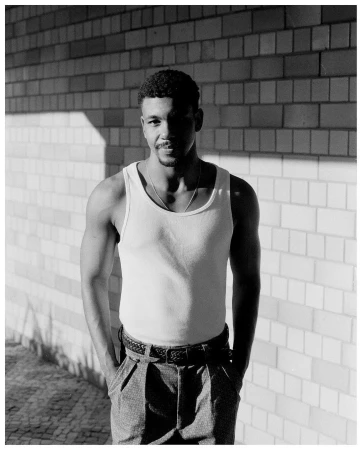 Portrait of a Black man in a white tank top standing against a brick wall, photographed by Rodrigo Oliveira.