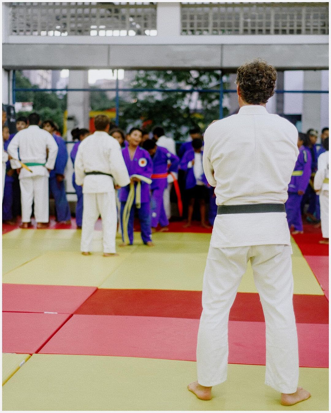Rear view of a man in judo uniform with folded arms in front of a group of judo athletes on a tatami mat in an indoor setting.