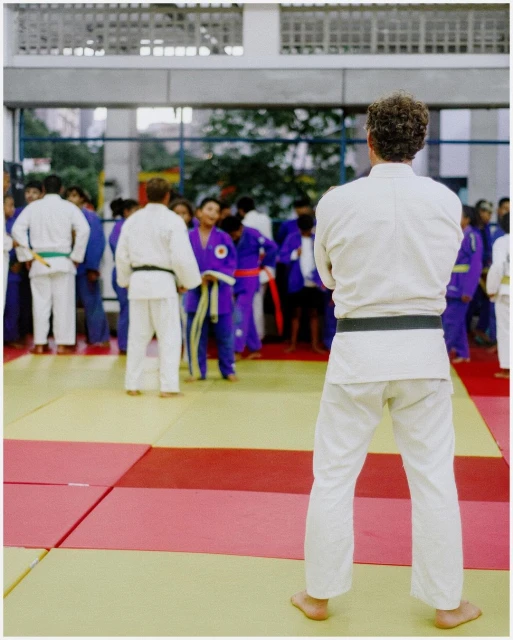 Rear view of a man in judo uniform with folded arms in front of a group of judo athletes on a tatami mat in an indoor setting.