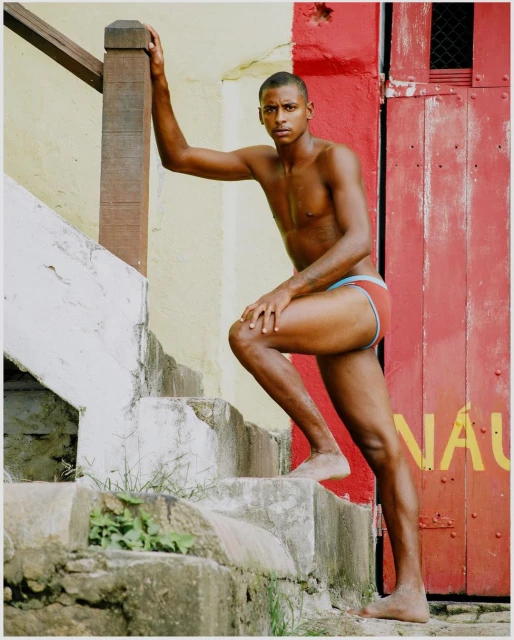 Athletic, dark-skinned man wearing swimwear, poses on concrete stairs with a red door and a yellow wall in the background.