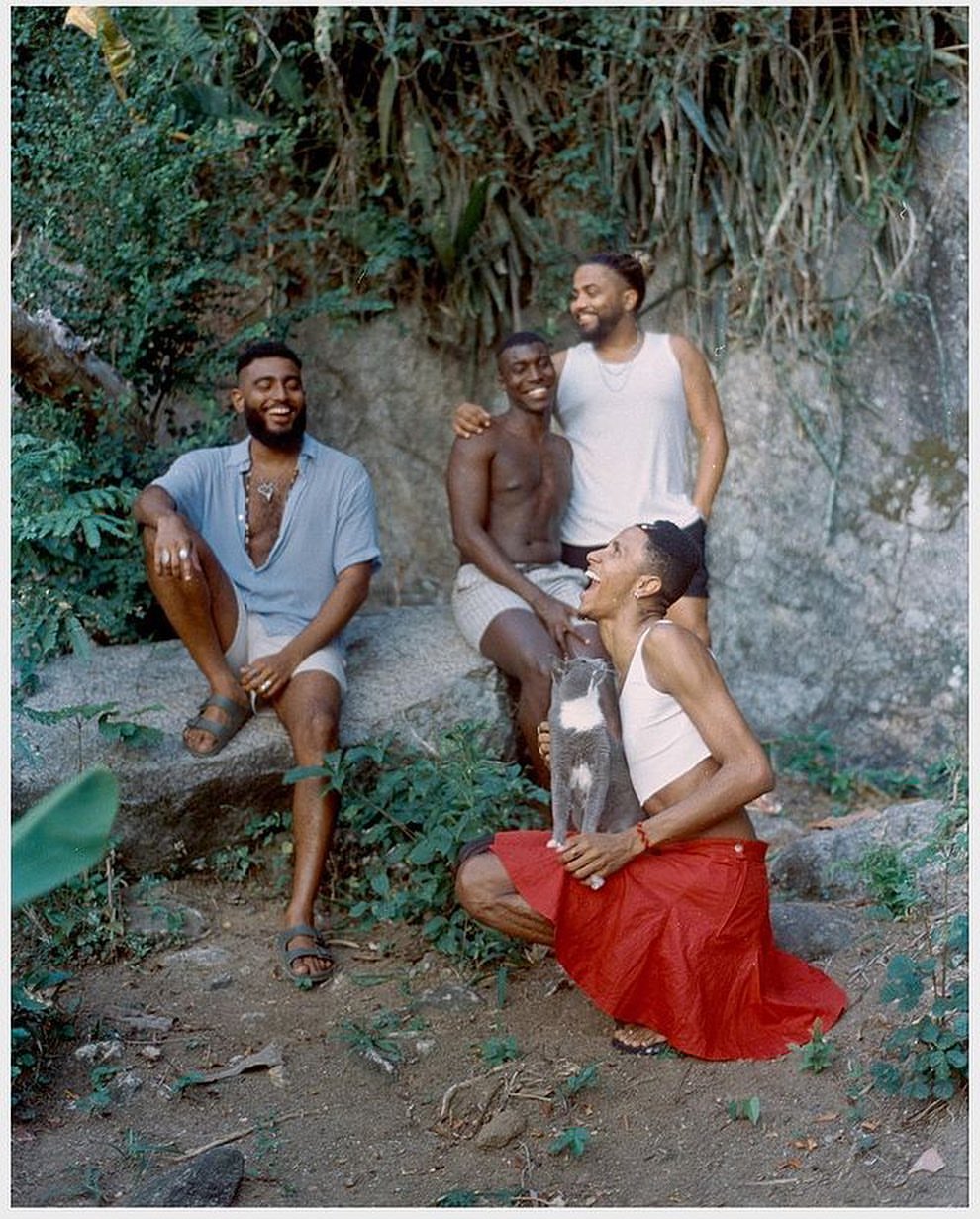 Four adult male friends laughing together in outdoor portrait with green foliage and rocks. One man holds a gray and white cat.