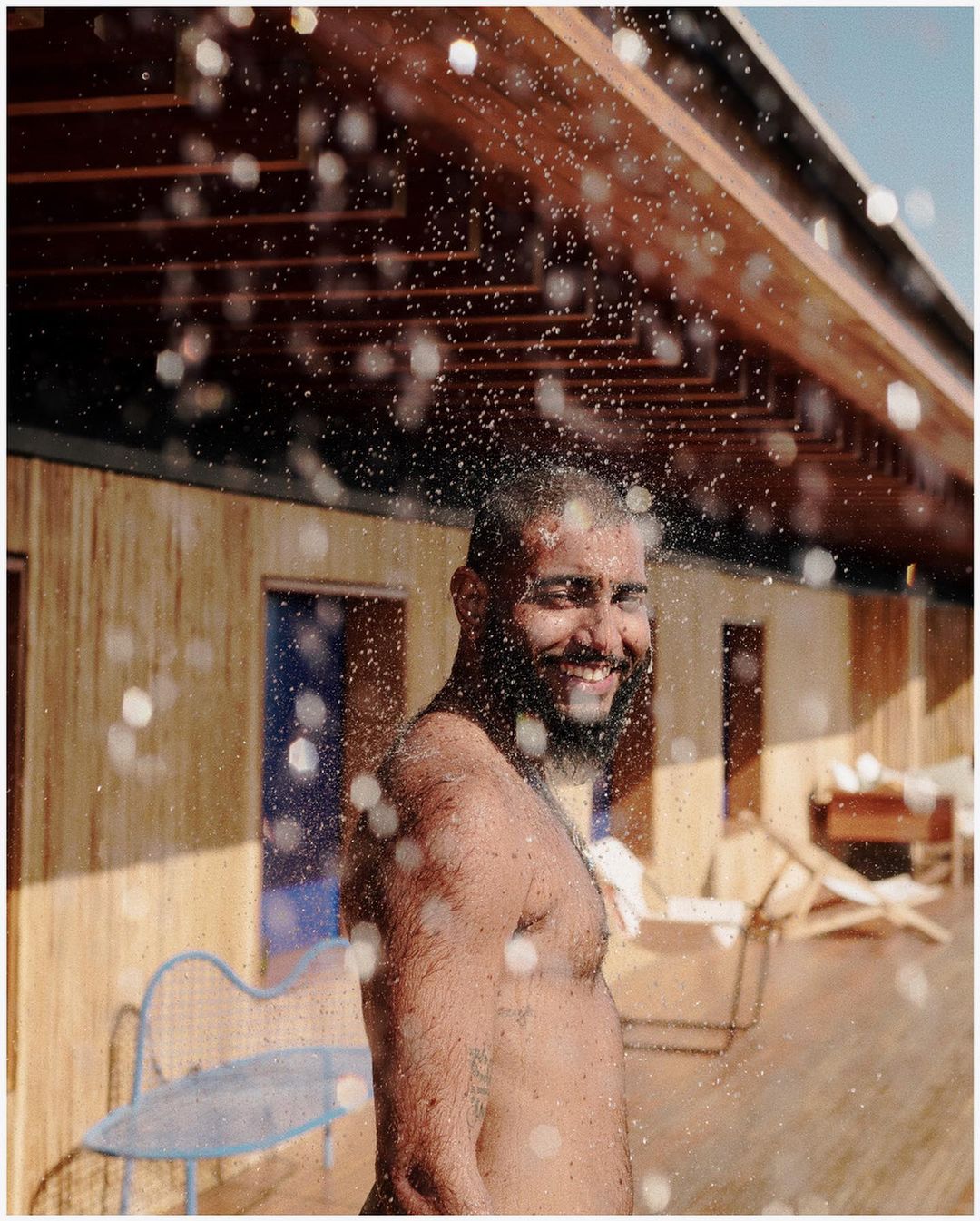 Adult male with beard smiles under outdoor shower, sunlight streams through roof beams in backyard setting, creating a refreshing summer vibe.