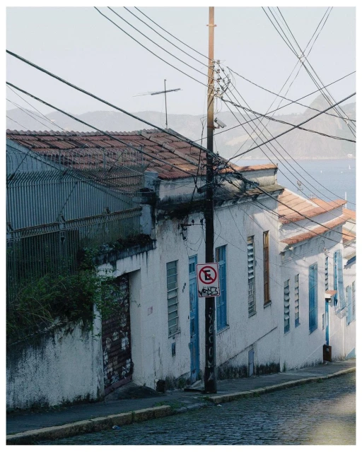 Urban landscape featuring weathered white buildings with terra cotta roofs, utility poles with wires, and a glimpse of water in the distance.