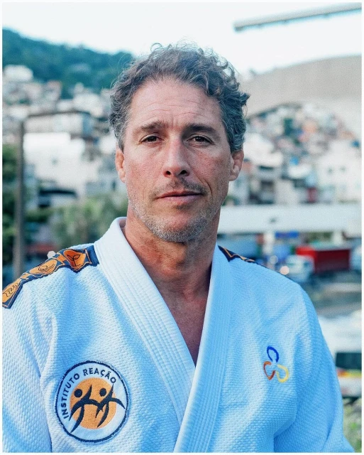Portrait of an adult man in a white martial arts uniform with logos, posing outdoors against a blurred urban background in daylight.